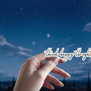 May include: A hand holds up a white sticker with the text "think happy thoughts" written in black cursive against a blue night sky with stars.