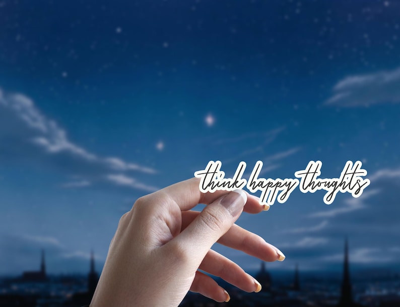 May include: A hand holds up a white sticker with the text "think happy thoughts" written in black cursive against a blue night sky with stars.