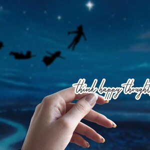 May include: A white sticker with the text "Think happy thoughts" in black script font. The sticker is held in a hand against a blue background with silhouettes of people flying.