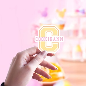 May include: A yellow and white sticker in the shape of the letter "C" with the name "COOKIEANN" in pink. The sticker is held in front of a blurred pink background with desserts.