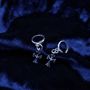 May include: A pair of silver-toned hoop earrings featuring dangling cross charms. Each cross charm is detailed with a small, central clear stone accent. The earrings are constructed with a hinged closure mechanism for secure attachment.