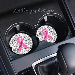 May include: Two pink and white car coasters with the words "Believe", "Warrior", "Strong", "Fighter", "Survivor", "Love", "Hope", and "Believe" repeated in a circle around a pink ribbon. The coasters are in a car cup holder.
