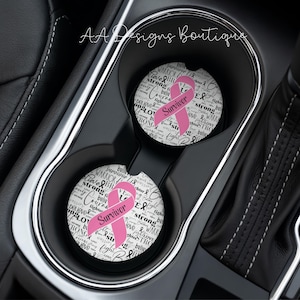 May include: Two circular car cup holder coasters featuring a white background with black typography including words like warrior, strong, and believe. Each coaster displays a prominent pink ribbon with the text Survivor printed across it.