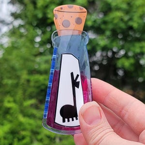 May include: A clear, bottle-shaped acrylic pin with a cork stopper. The bottle contains a purple liquid and a white graphic of a llama and musical note. The pin is held in a hand against a blurred green background.