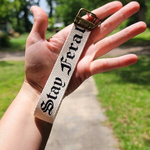 May include: A cream-colored keychain strap with the words "Stay Feral" in black script. The strap has a brass-colored metal clasp. The background is a blurred outdoor scene with greenery and a pathway.