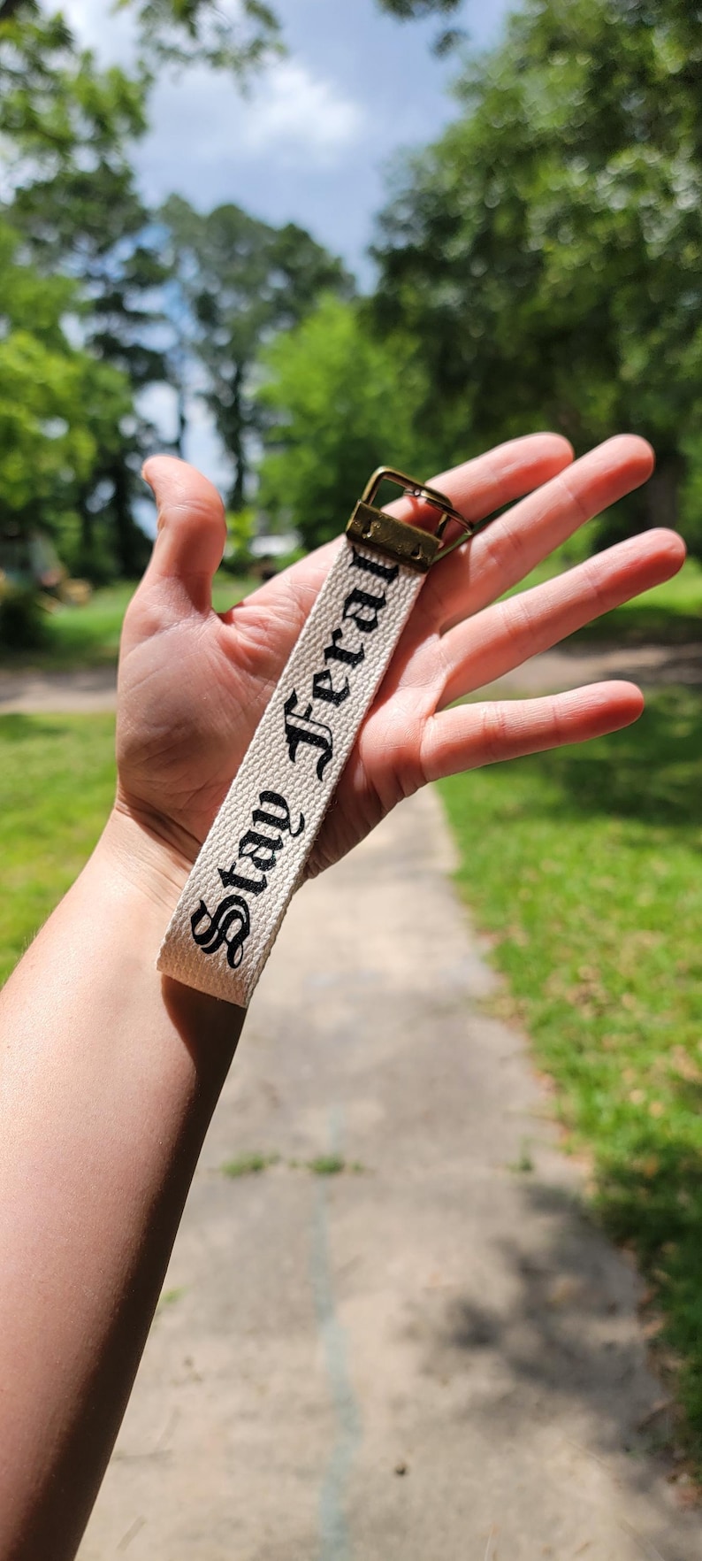 May include: A cream-colored keychain strap with the words "Stay Feral" in black script. The strap has a brass-colored metal clasp. The background is a blurred outdoor scene with greenery and a pathway.