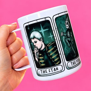 May include: A white ceramic mug featuring two rectangular tarot card illustrations. The left card shows a person in a military-style jacket labeled THE STAR. The right card depicts a skeleton figure in a uniform labeled THE FOOL.