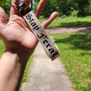 May include: A beige keychain strap with the words "Stay Feral" printed in black gothic lettering. The strap is attached to a metal clasp. The background is a blurred outdoor scene with trees and a pathway.