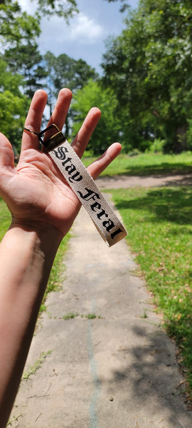 May include: A beige keychain strap with the words "Stay Feral" printed in black gothic lettering. The strap is attached to a metal clasp. The background is a blurred outdoor scene with trees and a pathway.