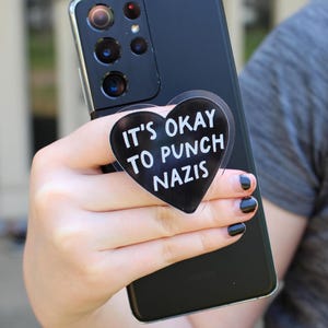 May include: A black smartphone with a heart-shaped phone grip attached. The grip has white text that reads "IT'S OKAY TO PUNCH NAZIS". The phone is held in a person's hand.