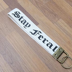 May include: White canvas keychain strap with the words "Stay Feral" printed in black gothic font. The strap has a bronze-colored metal clasp and ring. The keychain is lying on a wooden surface.
