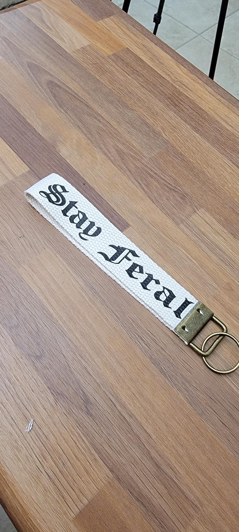 May include: White canvas keychain strap with the words "Stay Feral" printed in black gothic font. The strap has a bronze-colored metal clasp and ring. The keychain is lying on a wooden surface.