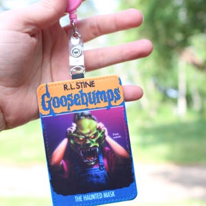 May include: A rectangular ID badge holder featuring the R.L. Stine Goosebumps The Haunted Mask book cover art, with a bright orange top section, a blue border, and a pink retractable reel attached to a clear plastic strap at the top.