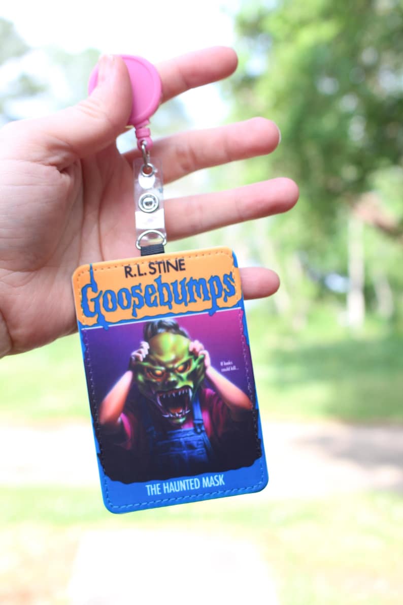 May include: A rectangular ID badge holder featuring the R.L. Stine Goosebumps The Haunted Mask book cover art, with a bright orange top section, a blue border, and a pink retractable reel attached to a clear plastic strap at the top.