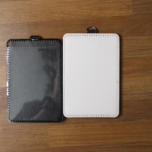 May include: Two rectangular badge holders with rounded corners and visible stitching along the edges. One holder is black with a clear plastic front window, and the other is solid white. Each holder features a small metal D-ring at the top center.