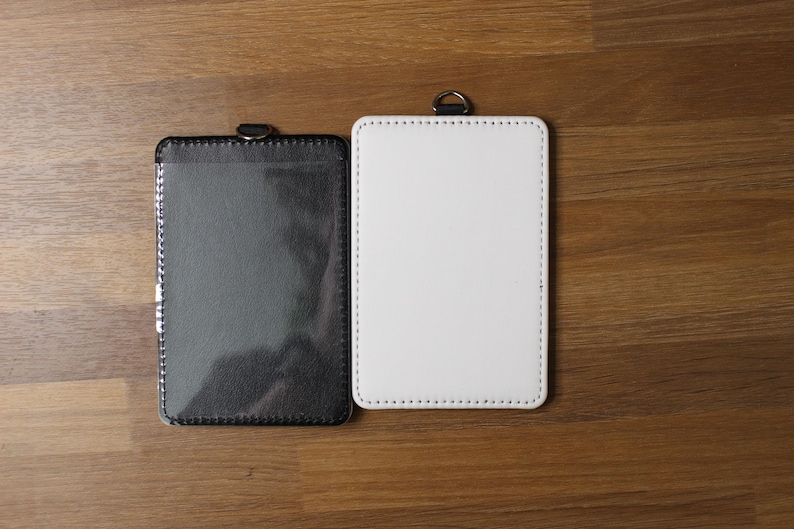 May include: Two rectangular badge holders with rounded corners and visible stitching along the edges. One holder is black with a clear plastic front window, and the other is solid white. Each holder features a small metal D-ring at the top center.