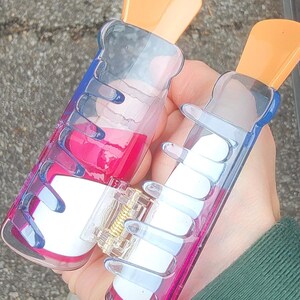 Op de afbeelding: Twee doorzichtige plastic haarclips met oranje bovenkanten en blauwe en roze accenten. E&eacute;n clip bevat een roze vloeistof, de andere witte rechthoekige vormen. De clips worden in een hand gehouden.