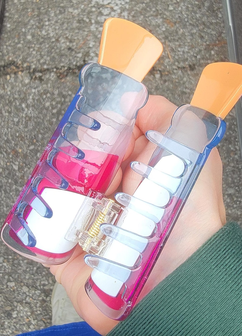 Op de afbeelding: Twee doorzichtige plastic haarclips met oranje bovenkanten en blauwe en roze accenten. E&eacute;n clip bevat een roze vloeistof, de andere witte rechthoekige vormen. De clips worden in een hand gehouden.