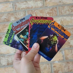 May include: A set of four rectangular Goosebumps book cover keychains featuring R.L. Stine branding. The designs include The Haunted Mask with a green monster, a blue house, a skeleton chef, and a dark forest scene, each with a metal ring attachment.