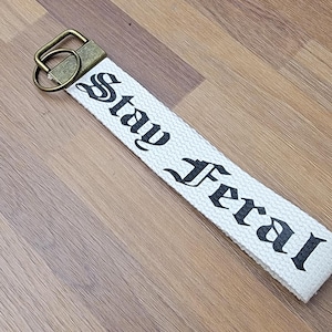 May include: White fabric keychain with the words "Stay Feral" printed in black gothic font. The keychain has a bronze-colored metal clasp and ring. The item is lying on a wooden surface.