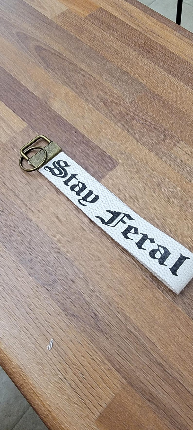 May include: White fabric keychain with the words "Stay Feral" printed in black gothic font. The keychain has a bronze-colored metal clasp and ring. The item is lying on a wooden surface.