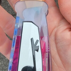 Op de afbeelding: Een doorzichtige, plastic flesvormige sleutelhanger met een lama-ontwerp. De sleutelhanger heeft een roze en blauwe vloeistof binnenin en een kaasvormige bovenkant. De lama is zwart en de kaasvormige bovenkant heeft grijze stippen.