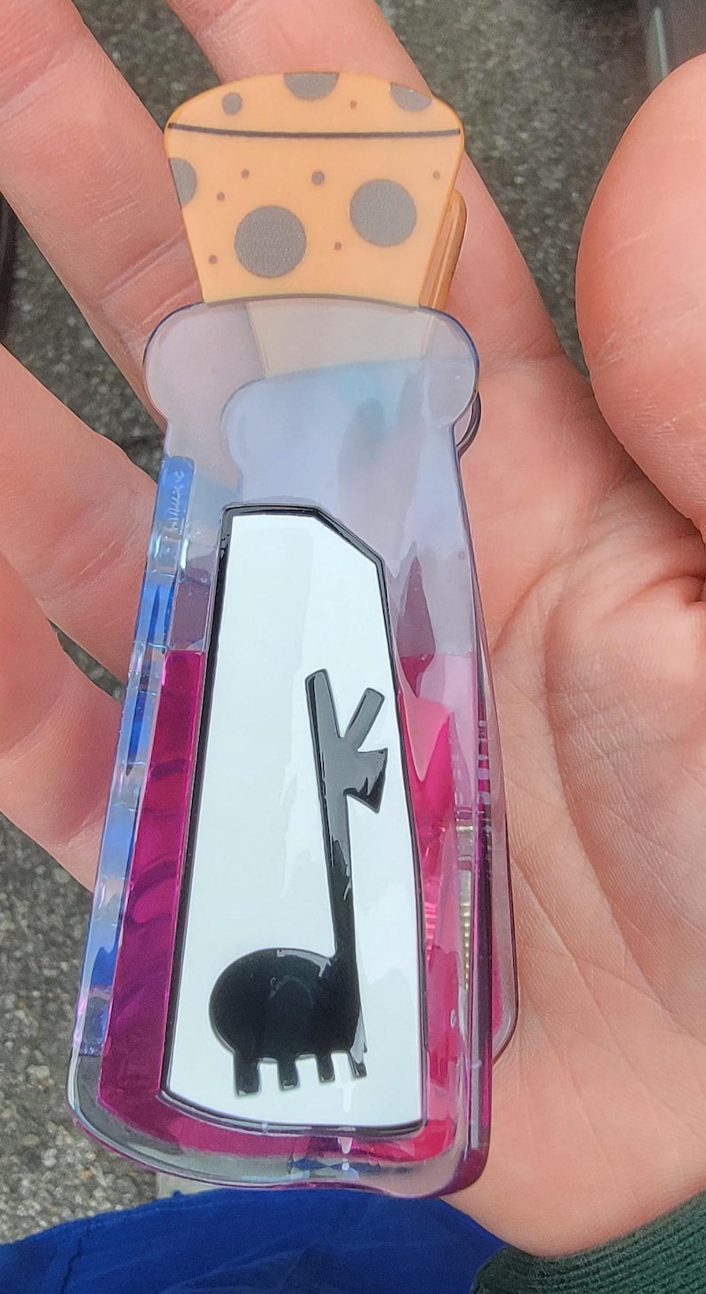Op de afbeelding: Een doorzichtige, plastic flesvormige sleutelhanger met een lama-ontwerp. De sleutelhanger heeft een roze en blauwe vloeistof binnenin en een kaasvormige bovenkant. De lama is zwart en de kaasvormige bovenkant heeft grijze stippen.