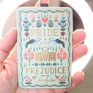 May include: A rectangular light green ID badge holder featuring a decorative illustration with the text Pride and Prejudice by Jane Austen. The design includes a central building, two birds, floral motifs, and a heart, finished with a metal ring.