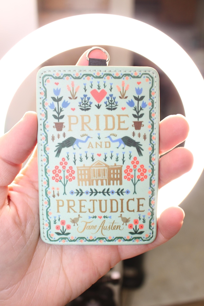 May include: A rectangular light green ID badge holder featuring a decorative illustration with the text Pride and Prejudice by Jane Austen. The design includes a central building, two birds, floral motifs, and a heart, finished with a metal ring.