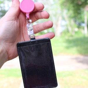 May include: A black rectangular badge holder with a clear plastic front window, attached to a pink circular retractable reel with a clear plastic strap and a metal snap button closure, designed for holding identification cards or security passes.
