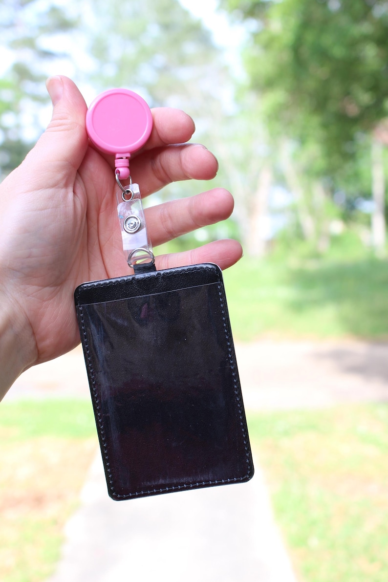 May include: A black rectangular badge holder with a clear plastic front window, attached to a pink circular retractable reel with a clear plastic strap and a metal snap button closure, designed for holding identification cards or security passes.