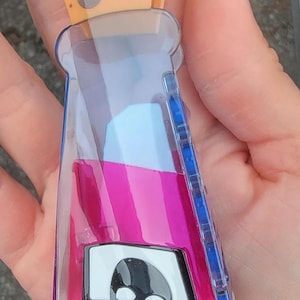 Op de afbeelding: Een doorzichtige, plastic flesvormige clip met een roze en blauw vloeistofontwerp en een kaasvormige bovenkant. De fles heeft een schedelafbeelding op de voorkant. De clip wordt in een hand gehouden.