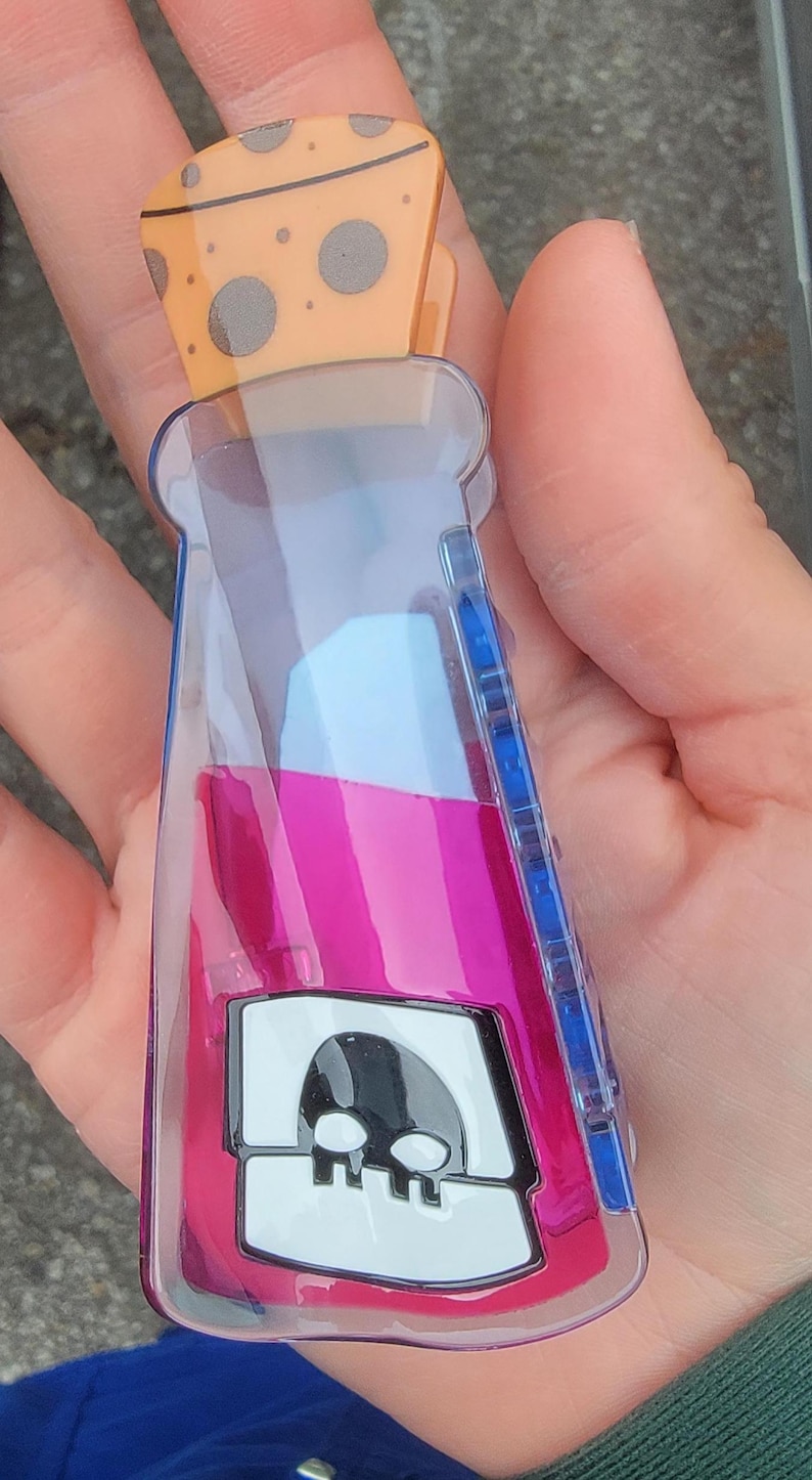 Op de afbeelding: Een doorzichtige, plastic flesvormige clip met een roze en blauw vloeistofontwerp en een kaasvormige bovenkant. De fles heeft een schedelafbeelding op de voorkant. De clip wordt in een hand gehouden.