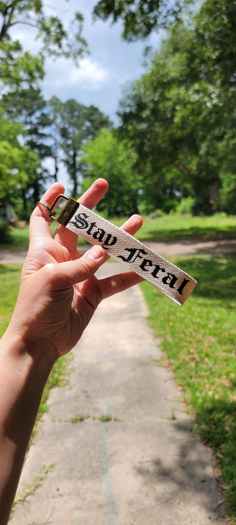 May include: A beige keychain strap with the words "Stay Feral" in black gothic lettering. The strap is attached to a brass-colored metal clasp. The background is a blurred outdoor scene with trees and a sidewalk.