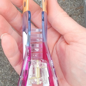 Op de afbeelding: Een doorzichtige en roze haarclip met een goudkleurig veermechanisme. De clip heeft een rechthoekige vorm met een gebogen ontwerp en wordt in een hand gehouden. De bovenkant van de clip is bruin met bruine vlekken.
