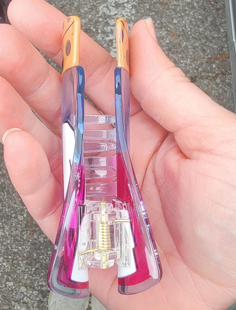 Op de afbeelding: Een doorzichtige en roze haarclip met een goudkleurig veermechanisme. De clip heeft een rechthoekige vorm met een gebogen ontwerp en wordt in een hand gehouden. De bovenkant van de clip is bruin met bruine vlekken.