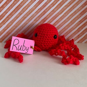 May include: A handmade red crochet octopus with black eyes and eight tentacles. A pink tag with the name "Ruby" is attached. The background features a white and brown striped pattern.