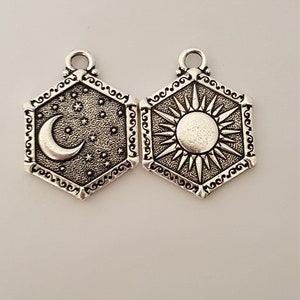 May include: Two silver charm pendants, one with a crescent moon and stars, the other with a sun with rays. Both pendants are hexagonal with ornate borders.