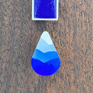May include: A rectangular watercolour paint pan filled with deep blue pigment and a teardrop-shaped colour swatch featuring a gradient of three distinct blue shades, ranging from a light sky blue at the top to a vibrant royal blue at the bottom.
