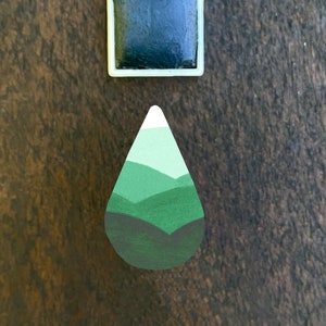 Könnte beinhalten: Zwei handgefertigte Magnete auf einer Holzoberfläche. Einer ist quadratisch mit einem dunkelgrünen Zentrum und einem weißen Rand. Der andere ist tropfenförmig mit einem Berglandschaftsdesign in Grün- und Weißtönen.