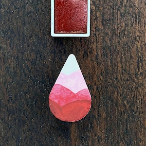 May include: A square watercolor paint pan filled with a deep red pigment, next to a teardrop-shaped painted design with shades of red, pink, and white. The items are on a dark wood surface.