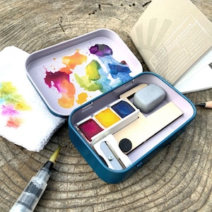 Pode incluir: Um kit de pintura em aquarela com uma lata azul-petróleo contendo tinta, um lápis, um pequeno caderno de esboços, um pincel de água e um pequeno pano branco com respingos de tinta coloridos. A tampa da lata tem uma paleta de aquarela.