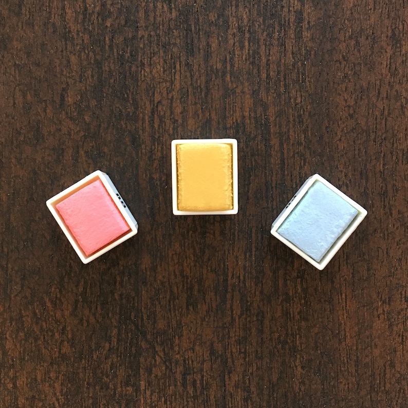 May include: Three watercolor paint pans, one pink, one yellow, and one light blue.