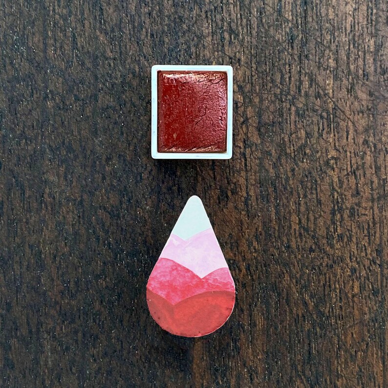May include: A square watercolor paint pan filled with a deep red pigment, next to a teardrop-shaped painted design with shades of red, pink, and white. The items are on a dark wood surface.