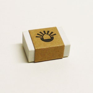 Puede incluir: Una pequeña caja blanca con una etiqueta de papel marrón. La etiqueta tiene un gráfico negro de un sol con una casa estilizada en su interior.