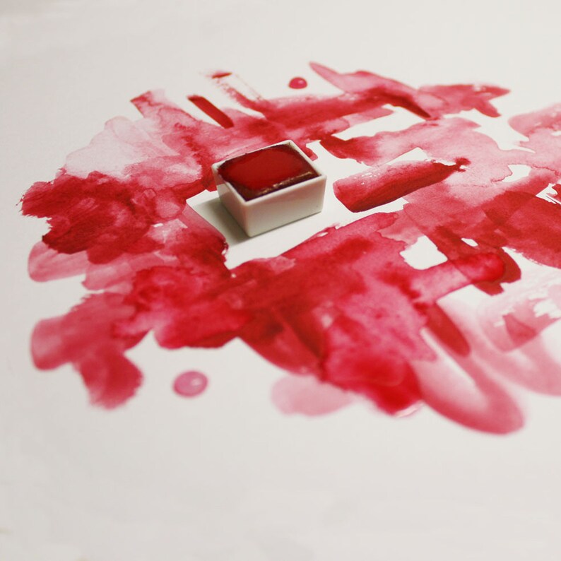 May include: A square, white watercolor paint pan filled with a vibrant red pigment sits on a white surface. The paint is surrounded by red watercolor brushstrokes and droplets, creating an abstract design. The image highlights art supplies.