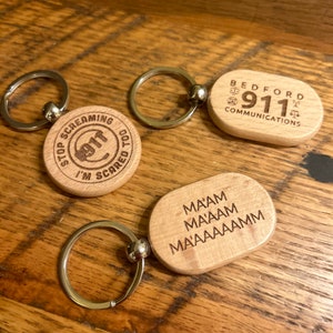May include: Wooden keychains with silver key rings. One keychain reads "STOP SCREAMING I'M SCARED TOO" around a 911 graphic. Another says "Bedford 911 Communications". The third keychain says "MA'AM MA'AM MA'AAAAAMM".