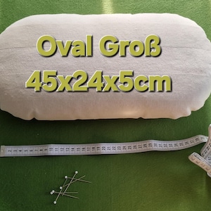 以下が含まれることがあります： オフホワイトの楕円形の枕で、黄色の文字で「Oval Groß 45x24x5cm」と書かれています。白いメジャーとピンが緑色の表面にあります。枕のサイズは約45 x 24 x 5 cmです。