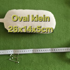 Puede incluir: Una herramienta de costura beige, de forma ovalada, con un asa, etiquetada como "Oval klein 26x14x5cm". Una cinta métrica blanca y alfileres también están sobre la superficie verde. La herramienta mide aproximadamente 26 x 14 x 5 cm.