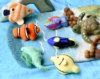 May include: A felt play set featuring a variety of colorful sea creatures, including a yellow fish, a blue fish, an orange and white fish, a green fish, a green sea turtle, and a brown and white whale. The set is on a blue felt background with a wooden archway.